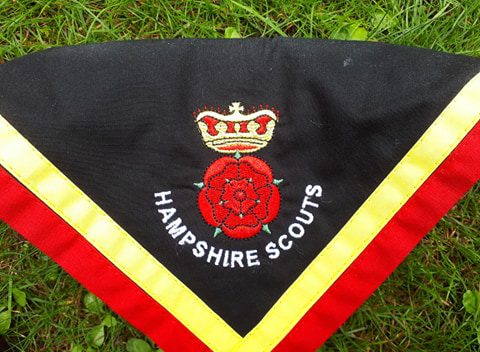 A Hampshire Scouts neckerchief with the Double rose and crown emblem embroidered on it. The scarf is black with gold and red trimming.