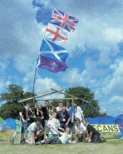 A group of Scouts sit underneath a constructed gateway with Union Flag, St. George's flag, St. Andrew's flag and WOSM flags.